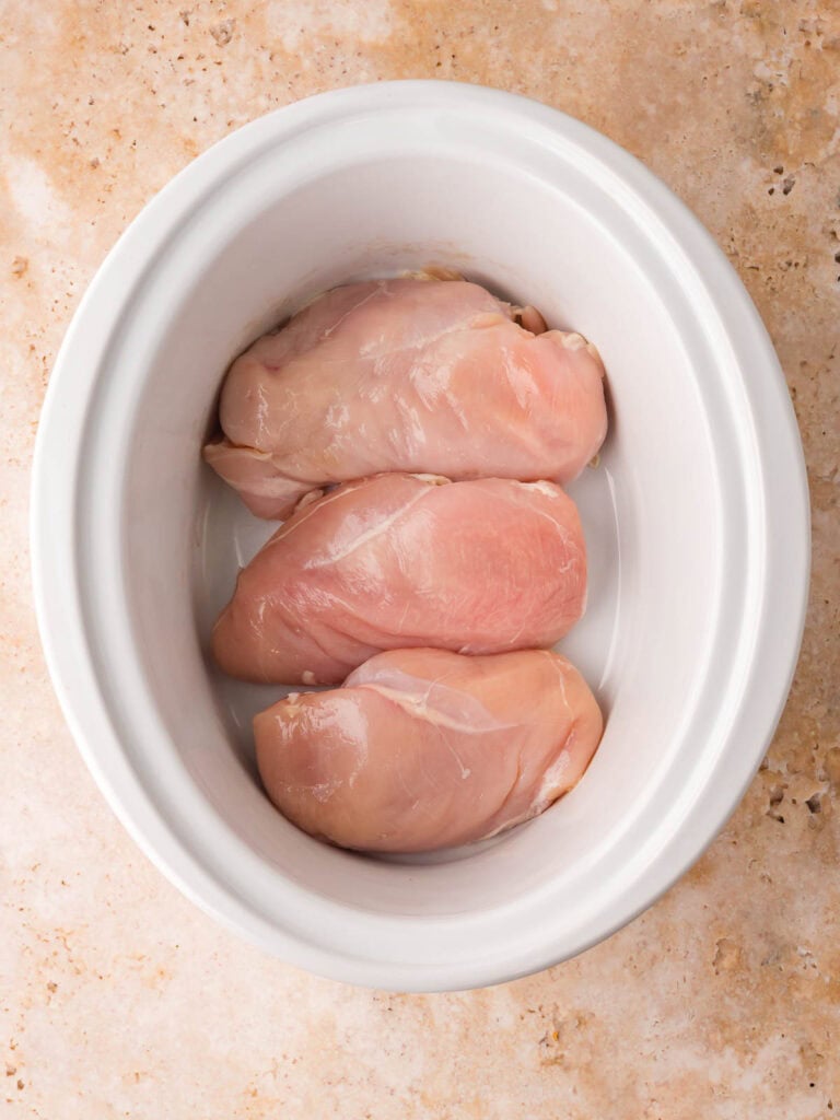 Three raw chicken breasts arranged in a white slow cooker for chicken and dumplings by Tessie's Table.