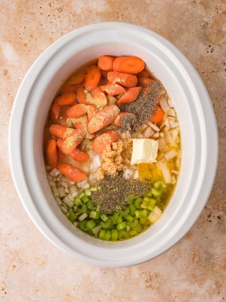 Sliced carrots, celery, onion, garlic, butter, and seasonings added to the slow cooker with broth by Tessie's Table.