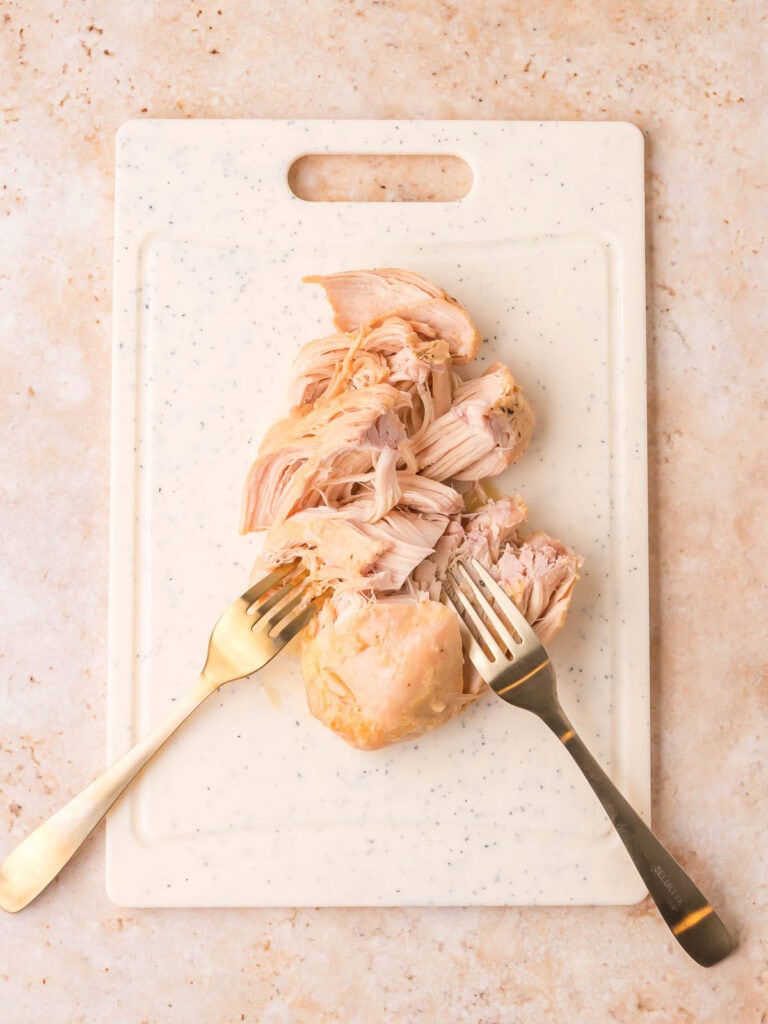 Cooked chicken breast being shredded with two forks on a cutting board by Tessie's Table.