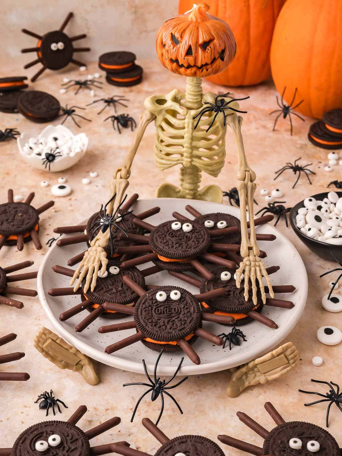 A plate of Oreo spider cookies held by a toy skeleton with pumpkins and candy eyes around it by Tessie's Table.
