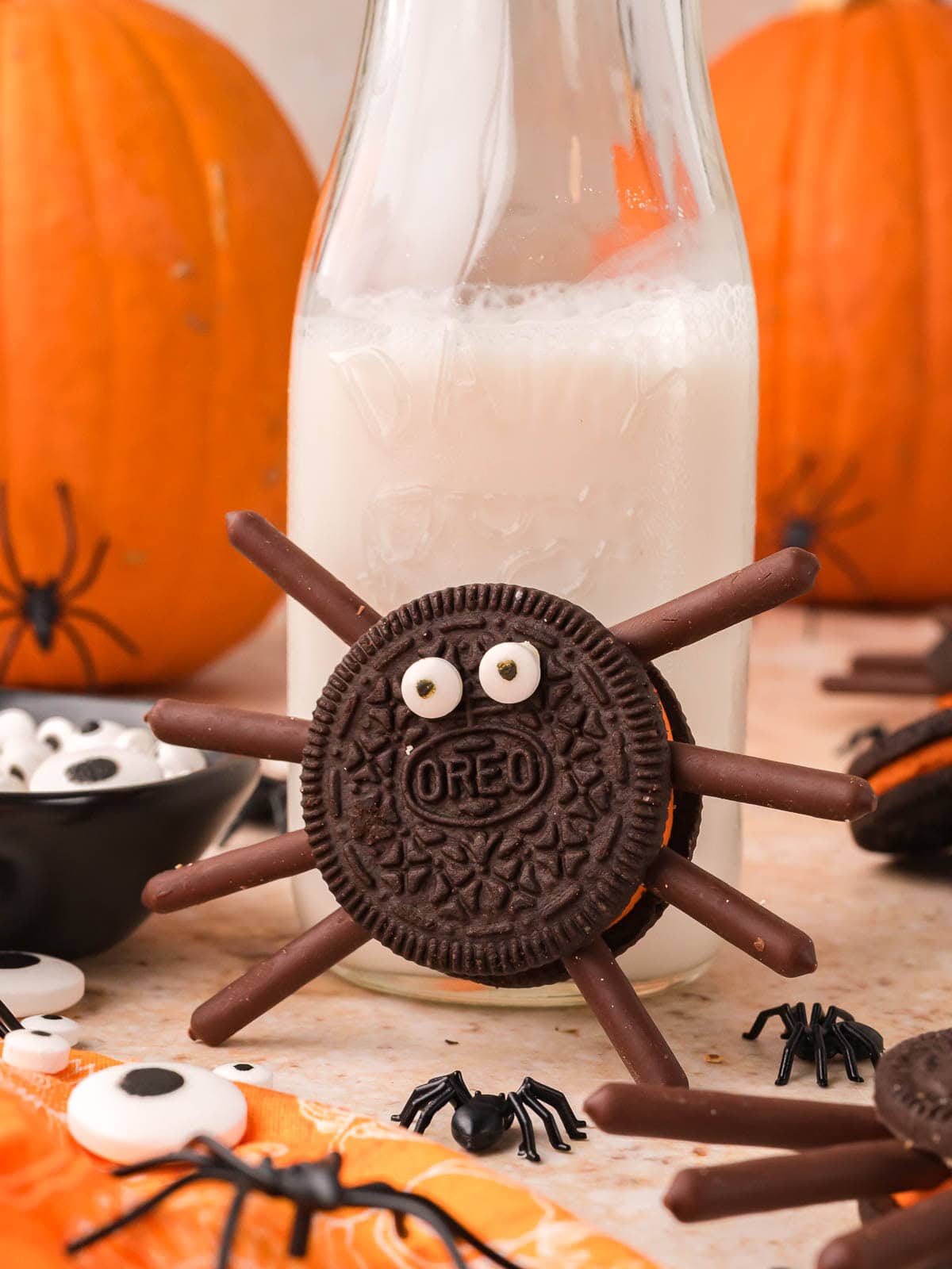 Oreo spider cookies with chocolate stick legs and candy eyes in front of a bottle of milk and pumpkins by Tessie's Table.