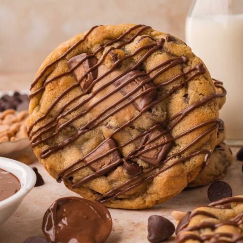 Nutella stuffed Reese's cookie with chocolate drizzle leaning against a glass of milk with a spoon of Nutella in front by Tessie's Table.
