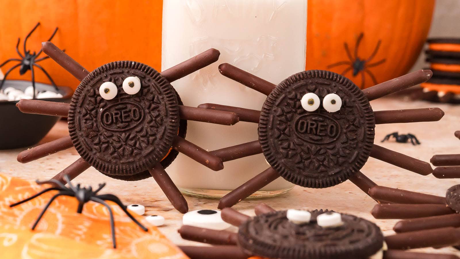 Two Oreo spider cookies with chocolate stick legs and candy eyes beside a bottle of milk and a pumpkin by Tessie's Table.