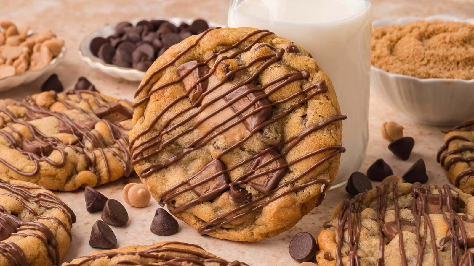 Nutella stuffed Reese's cookies with chocolate drizzle beside a glass of milk and bowls of chocolate chips and peanut butter chips by Tessie's Table.