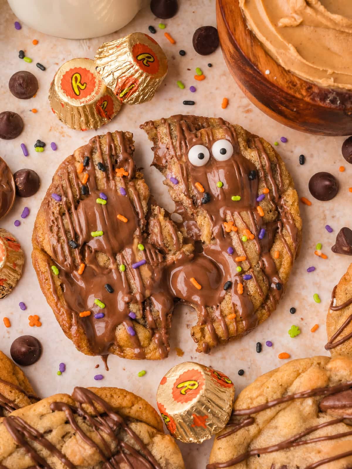 Halloween monster Nutella stuffed Reese's cookie broken in half with candy eyes, sprinkles and melted chocolate with Reese's cups beside it by Tessie's Table.