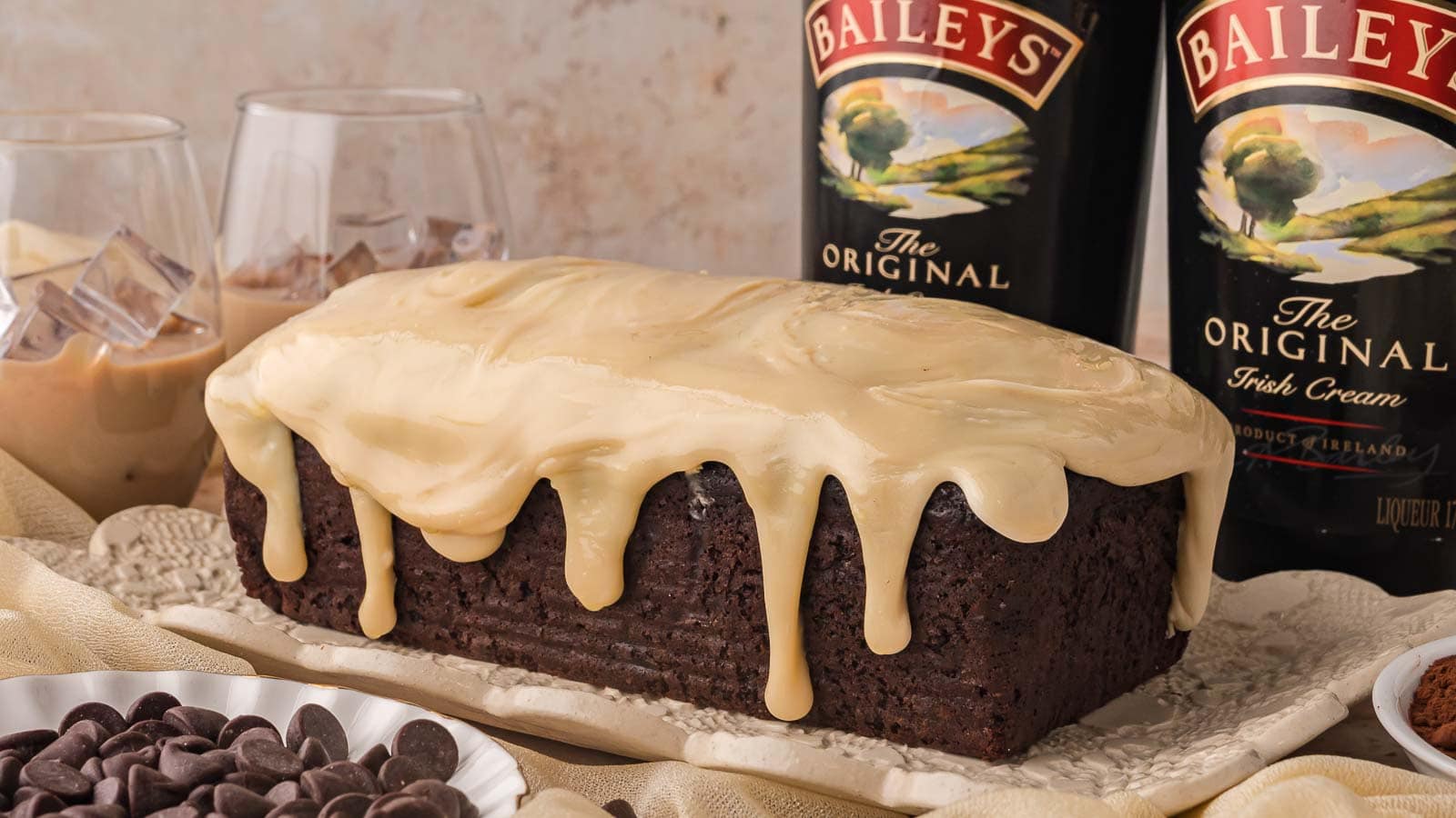 Chocolate Baileys banana bread loaf with white chocolate Baileys ganache dripping down the sides next to two bottles of Baileys Irish Cream by Tessie's Table.