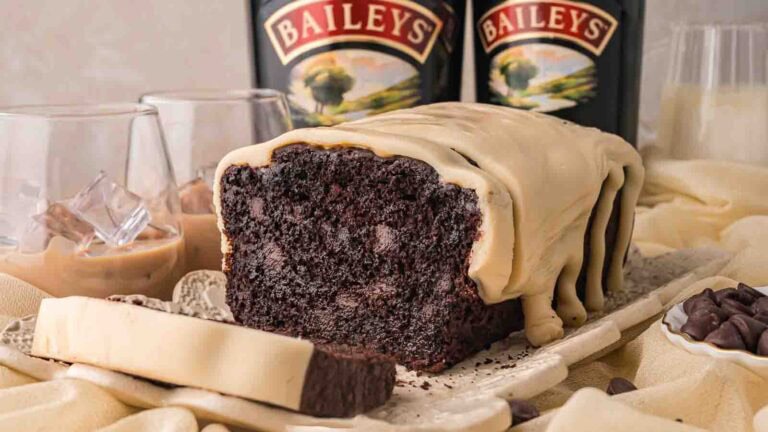Cut chocolate Baileys banana bread showing a dark chocolate crumb with chocolate chips under a white chocolate Baileys ganache, with Baileys bottles behind by Tessie's Table.