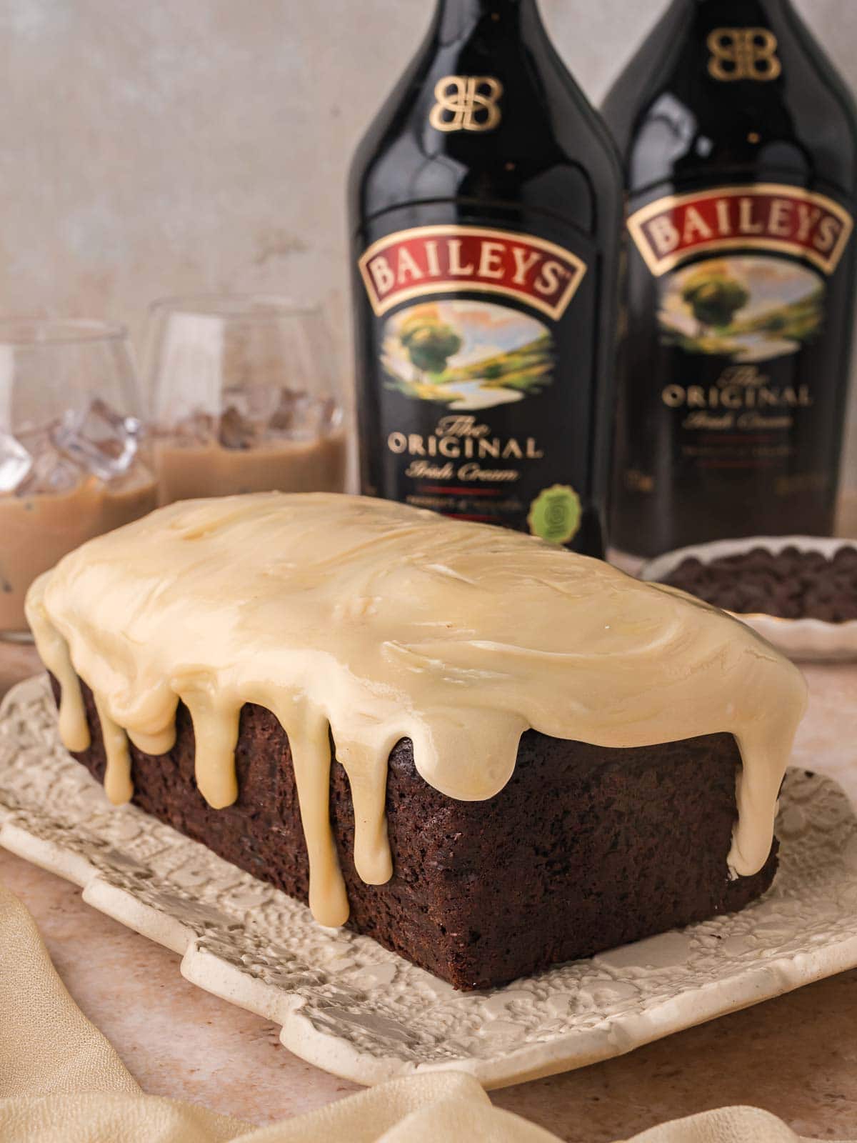 Whole chocolate Baileys banana bread loaf with white chocolate Baileys ganache dripping down the sides, with Baileys bottles and glasses of Baileys over ice behind by Tessie's Table.