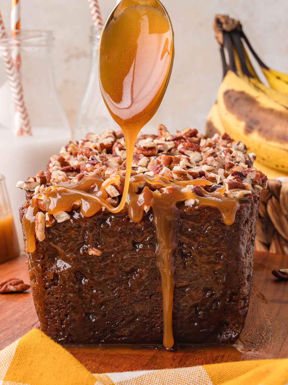 Caramel sauce being spooned over a caramel pecan banana bread loaf topped with chopped pecans, with ripe bananas and milk bottles behind it by Tessie's Table.