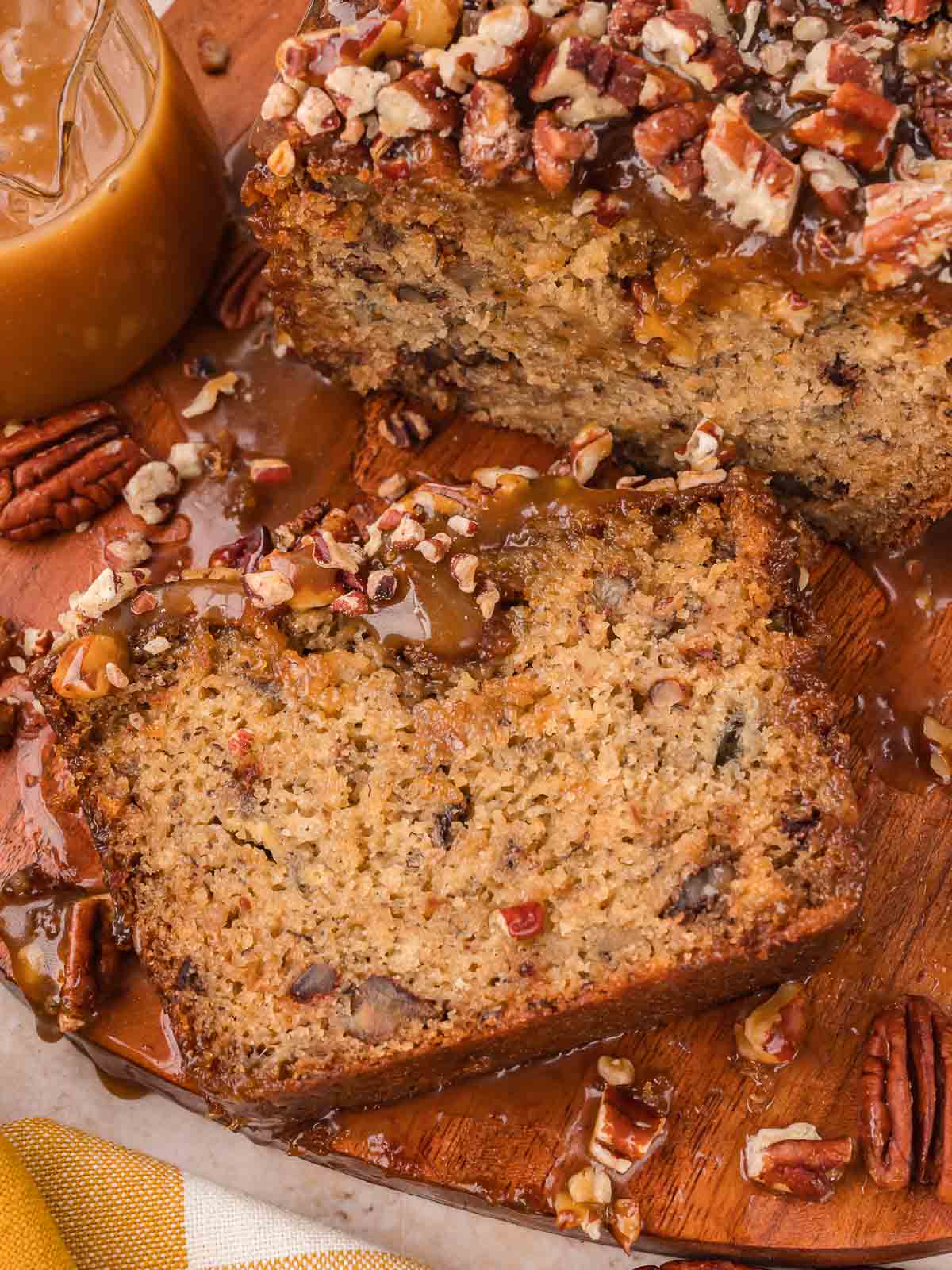 Slice of caramel pecan banana bread on a wooden board with caramel sauce and chopped pecans around it by Tessie's Table.