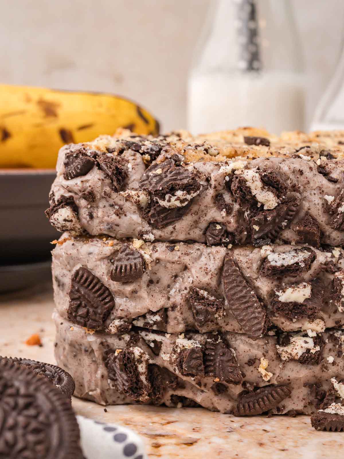 Stack of three Oreo banana bread slices covered in cookies and cream glaze and chopped Oreos with bananas and a bottle of milk behind by Tessie's Table.