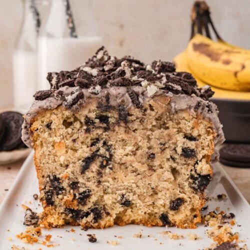 Cut end of an Oreo banana bread loaf on a white platter showing Oreo pieces through the crumb with milk bottles and bananas behind by Tessie's Table.