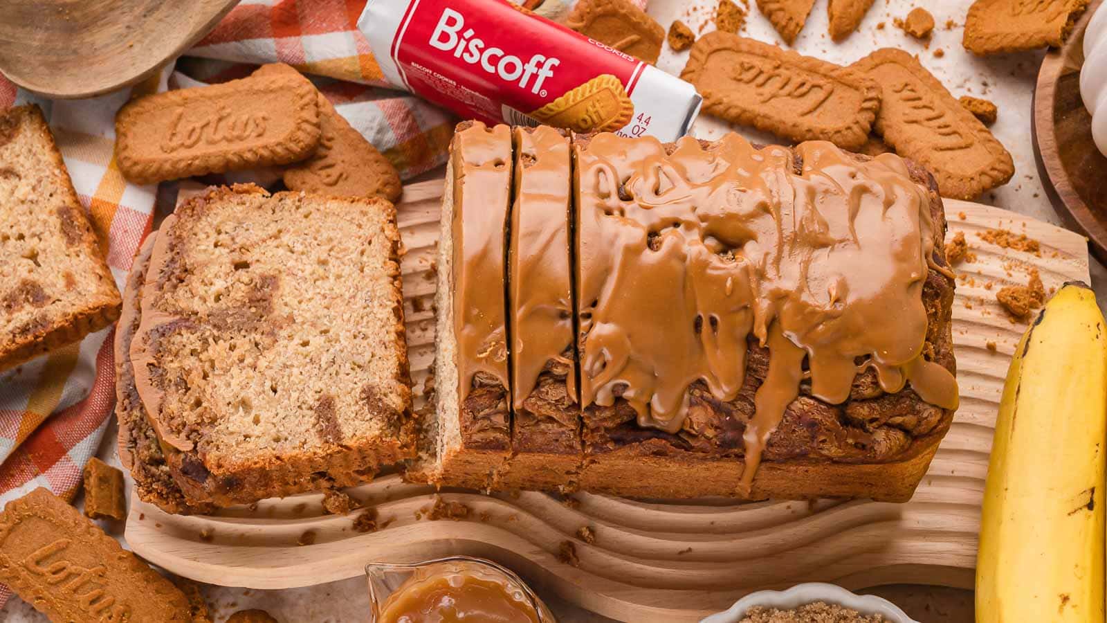 Overhead view of a sliced Biscoff banana bread loaf with cookie butter dripping over the top, surrounded by Lotus Biscoff cookies and a banana by Tessie's Table.