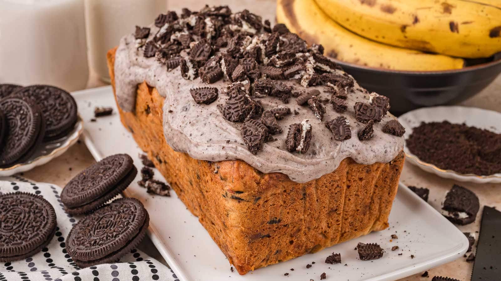 Whole Oreo banana bread loaf with thick cookies and cream glaze and chopped Oreos on top, with Oreos and ripe bananas around it by Tessie's Table.