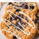 Blueberry crumble cookies with a white chocolate drizzle stacked on a white scalloped plate by Tessie's Table.