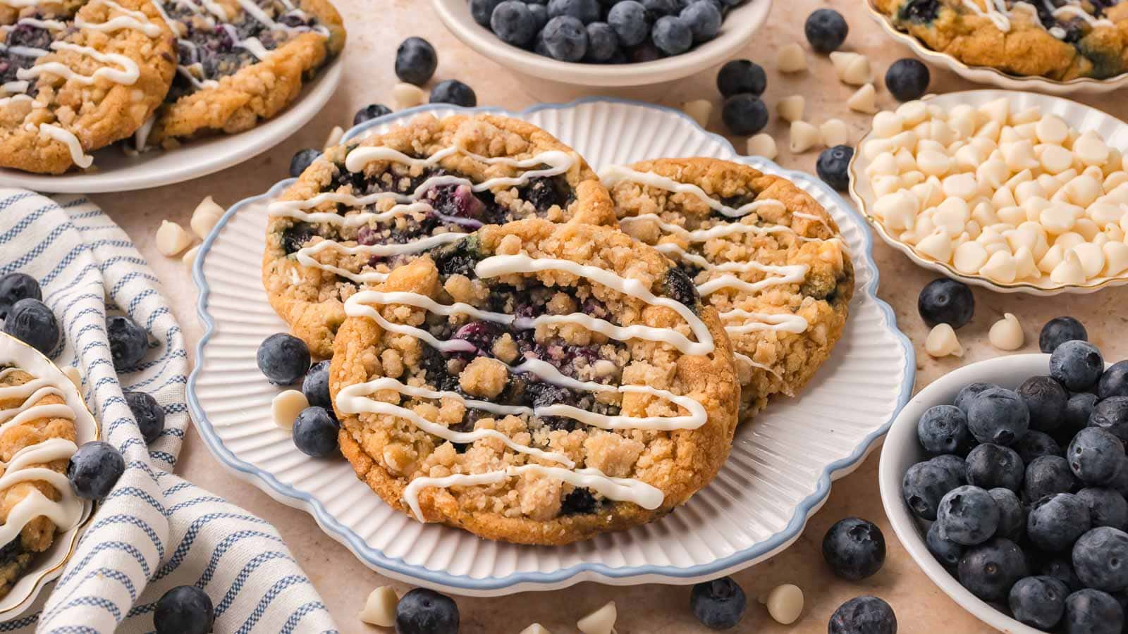 Blueberry crumble white chocolate cookies with a white chocolate drizzle on a white scalloped plate, surrounded by bowls of fresh blueberries and white chocolate chips by Tessie's Table.