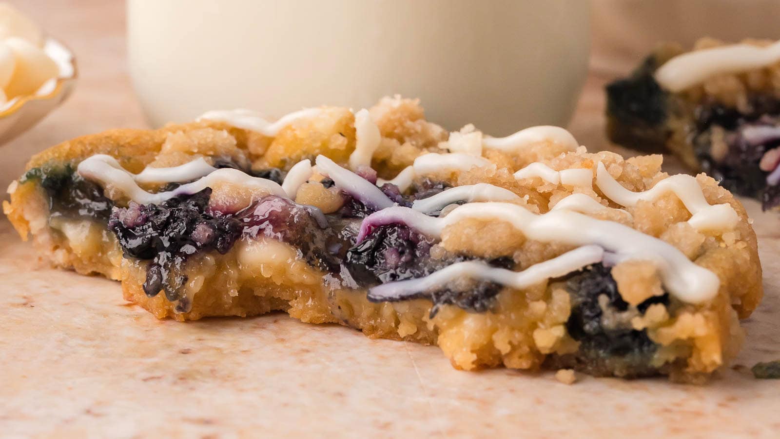 Blueberry crumble white chocolate cookie broken open to show the blueberry center, with crumble topping and a white chocolate drizzle, in front of a glass of milk by Tessie's Table.
