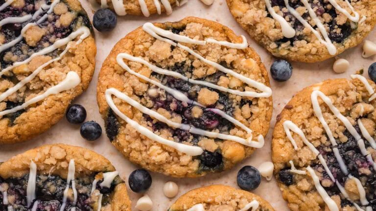 Overhead view of blueberry crumble white chocolate cookies with crumble topping and a white chocolate drizzle, scattered with fresh blueberries and white chocolate chips by Tessie's Table.