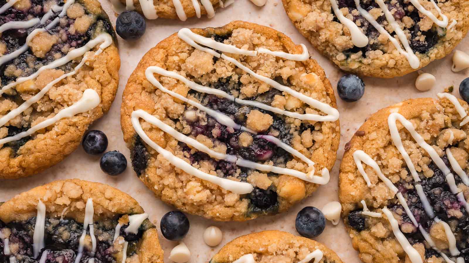 Overhead view of blueberry crumble white chocolate cookies with crumble topping and a white chocolate drizzle, scattered with fresh blueberries and white chocolate chips by Tessie's Table.