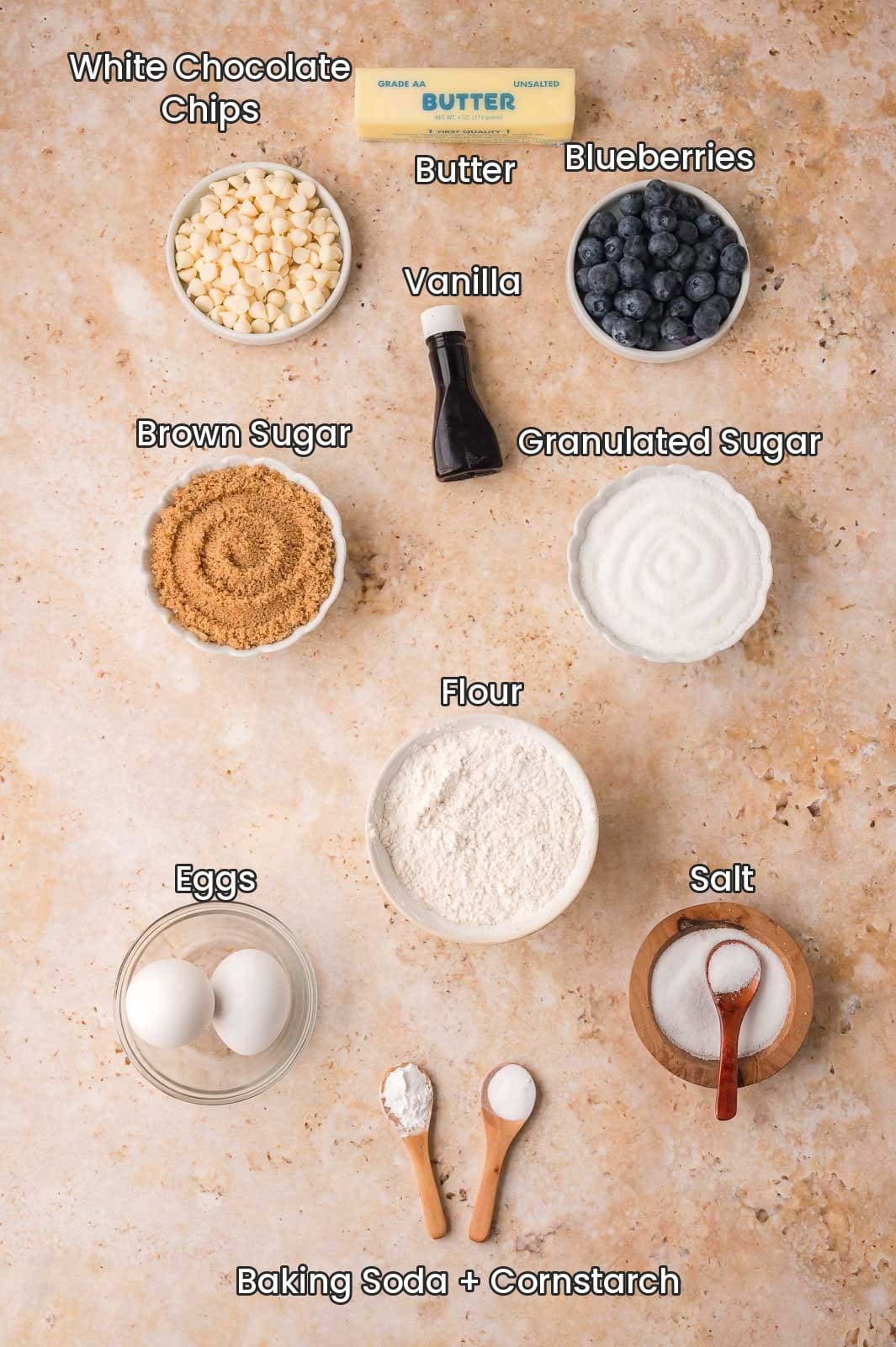Labeled ingredients for blueberry crumble white chocolate cookies including butter, white chocolate chips, fresh blueberries, vanilla, brown sugar, granulated sugar, flour, eggs, salt, baking soda and cornstarch by Tessie's Table.