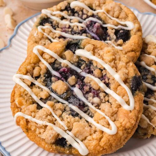 Two blueberry crumble white chocolate cookies on a white plate with a bowl of fresh blueberries behind by Tessie's Table.