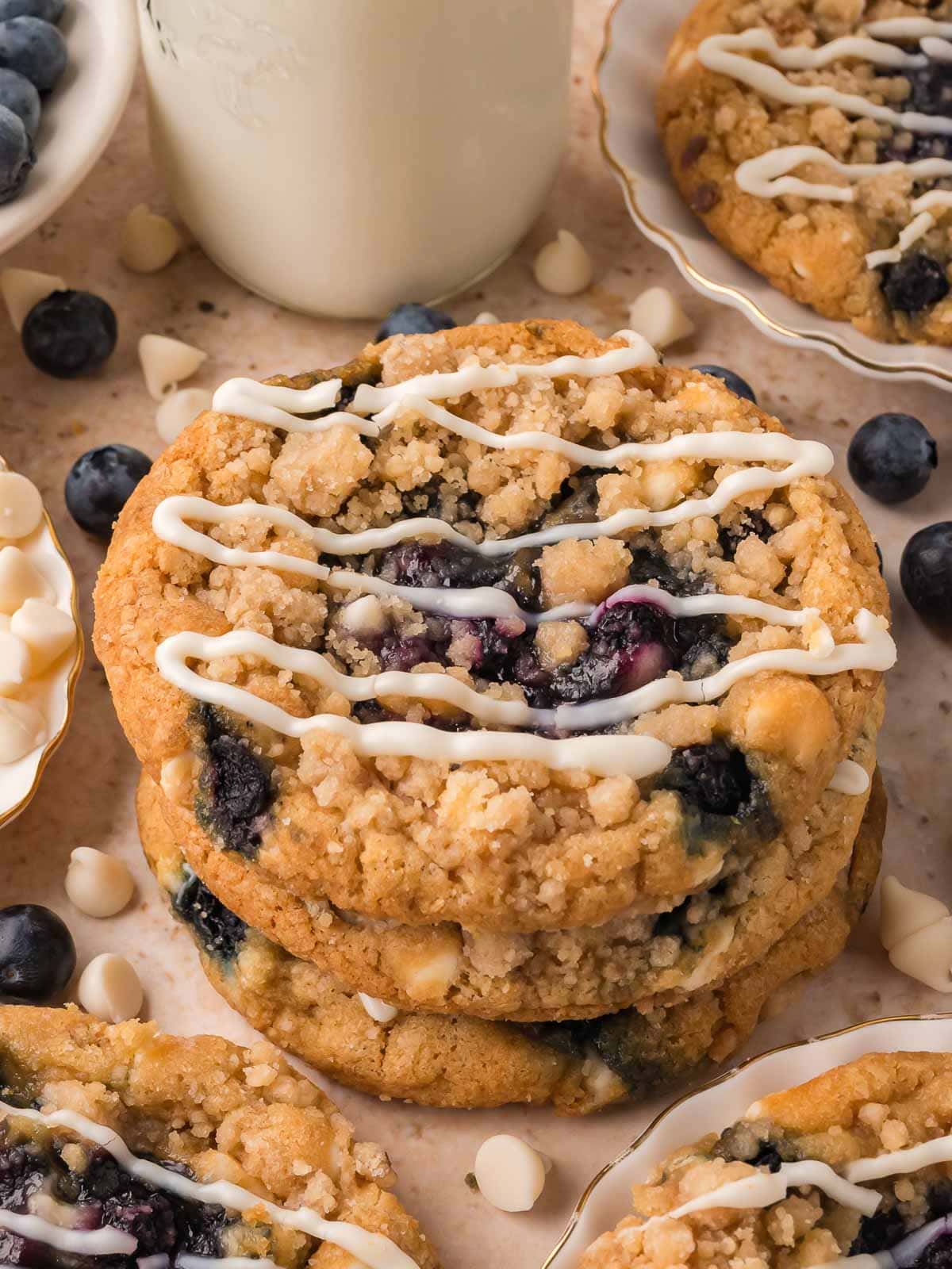 Stack of three blueberry crumble white chocolate cookies with a bottle of milk, fresh blueberries and white chocolate chips behind by Tessie's Table.