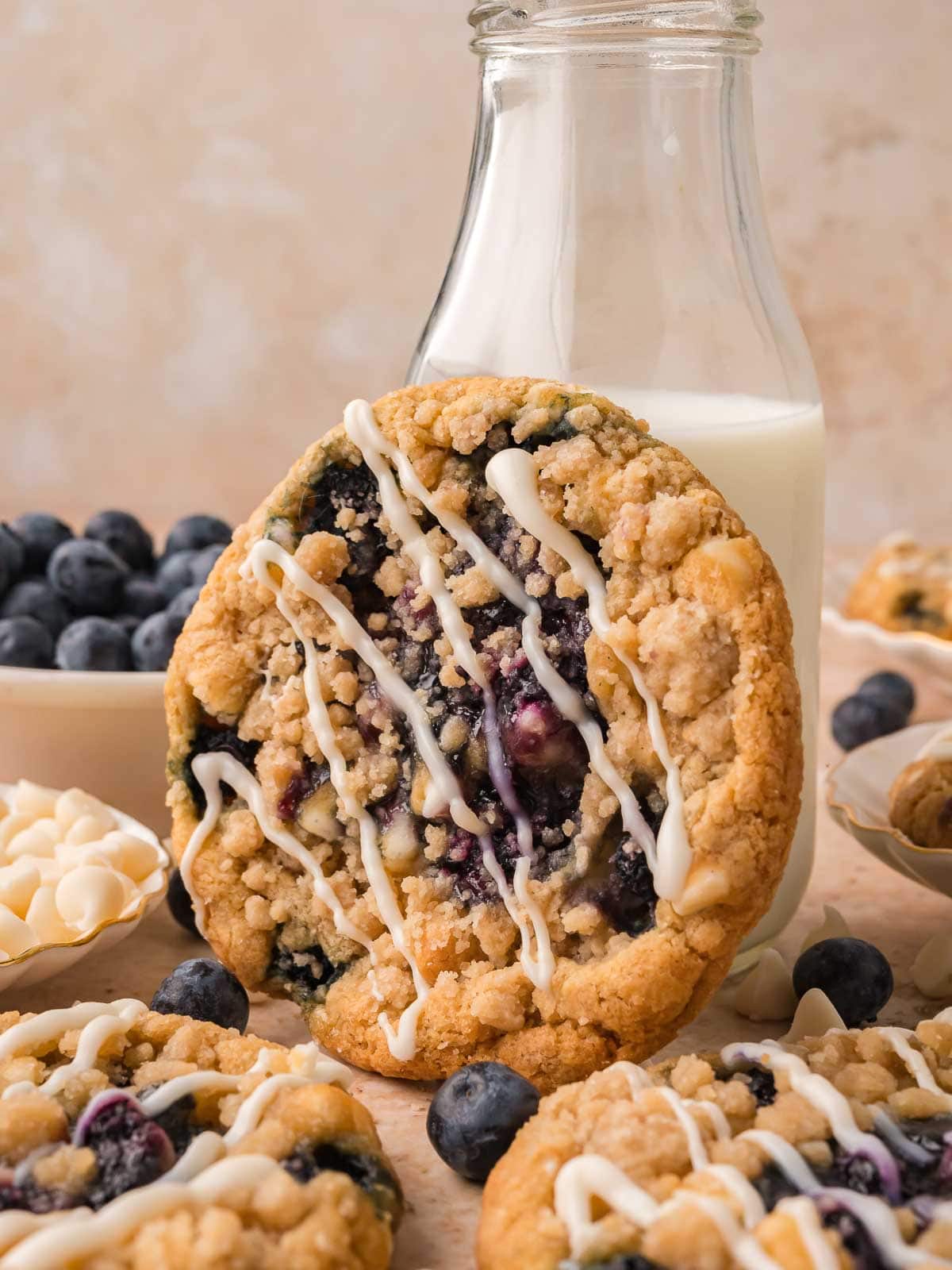 Blueberry crumble white chocolate cookie standing up in front of a bottle of milk with a bowl of fresh blueberries and more cookies around it by Tessie's Table.
