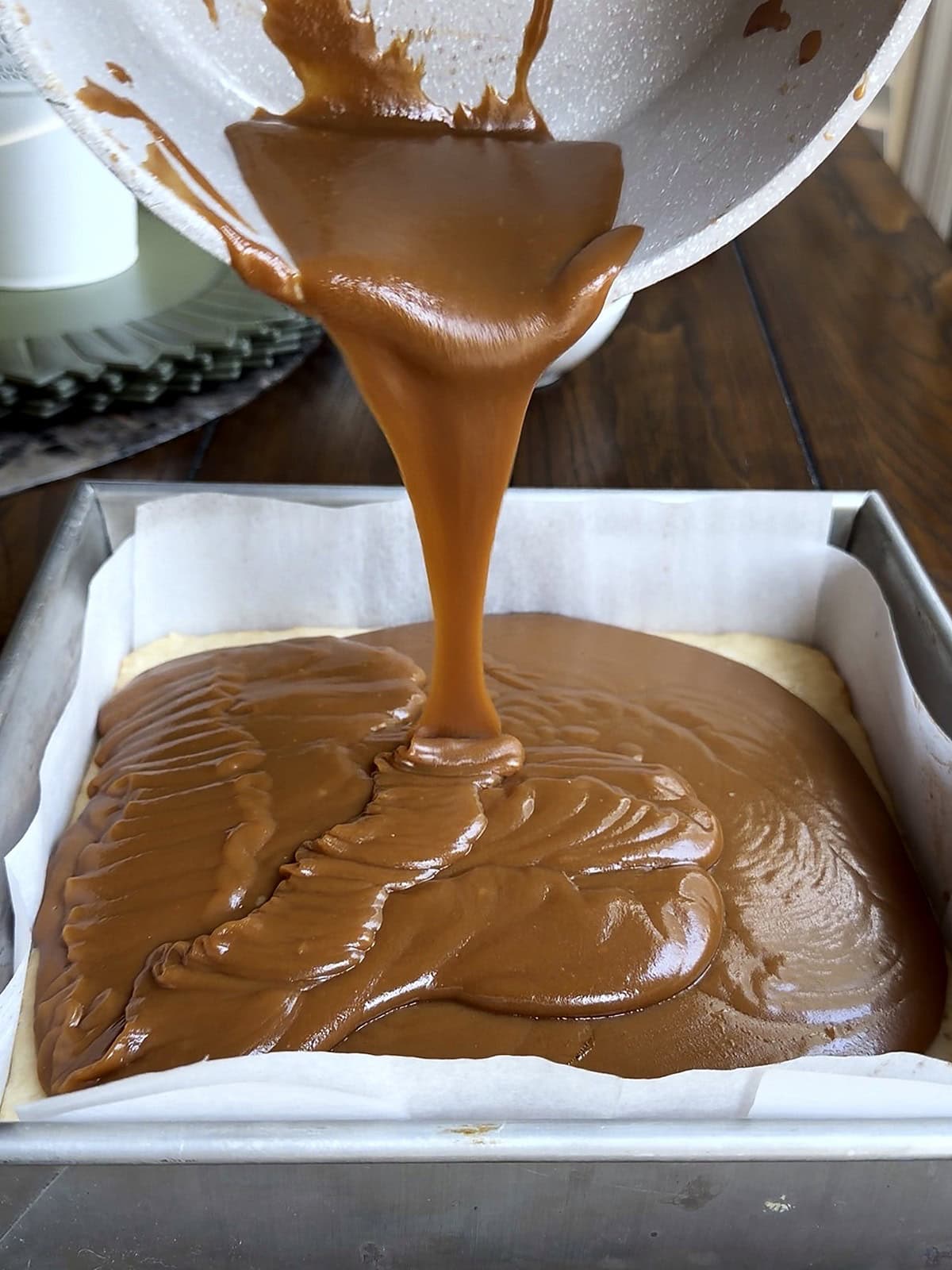 Homemade caramel with sweetened condensed milk being poured from a saucepan over a shortbread base in a lined square pan by Tessie's Table.