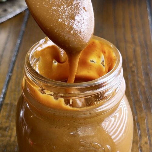 Spoon lifting thick homemade caramel with sweetened condensed milk out of a glass jar by Tessie's Table.