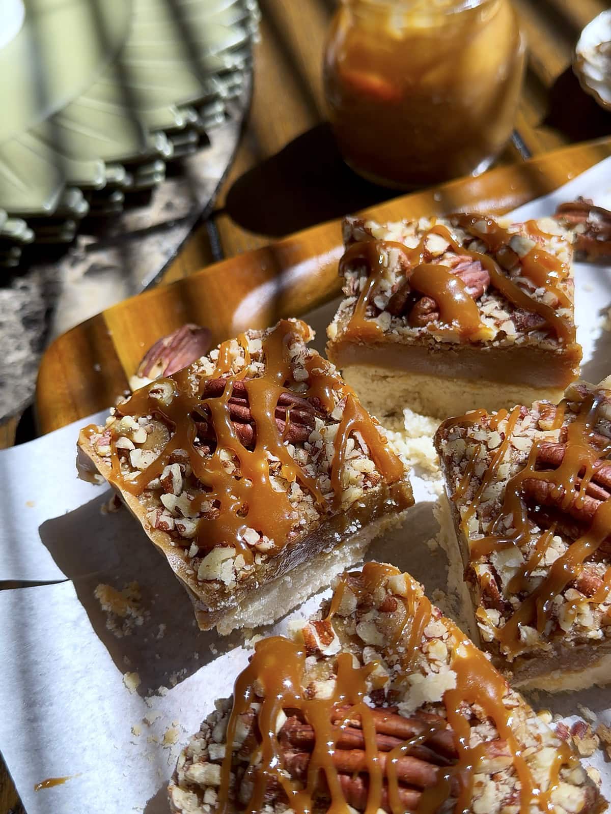 Salted caramel pecan bars on a shortbread base drizzled with homemade caramel, with a jar of caramel sauce behind by Tessie's Table.