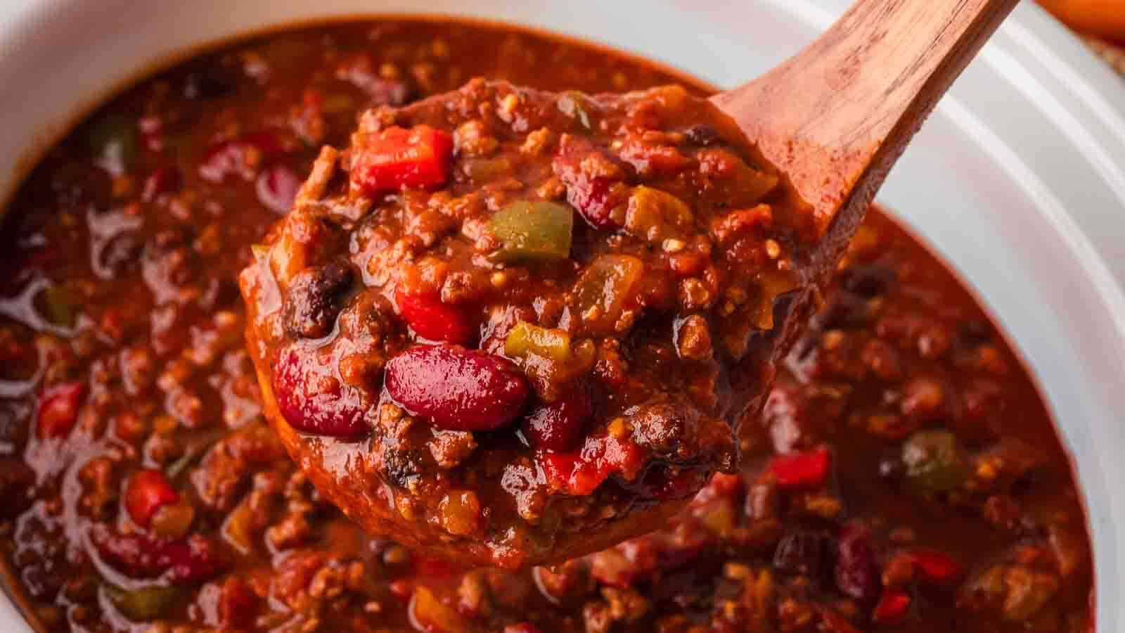 Wooden ladle lifting a scoop of slow cooker turkey chili with kidney beans, ground turkey and bell peppers out of the crockpot by Tessie's Table.