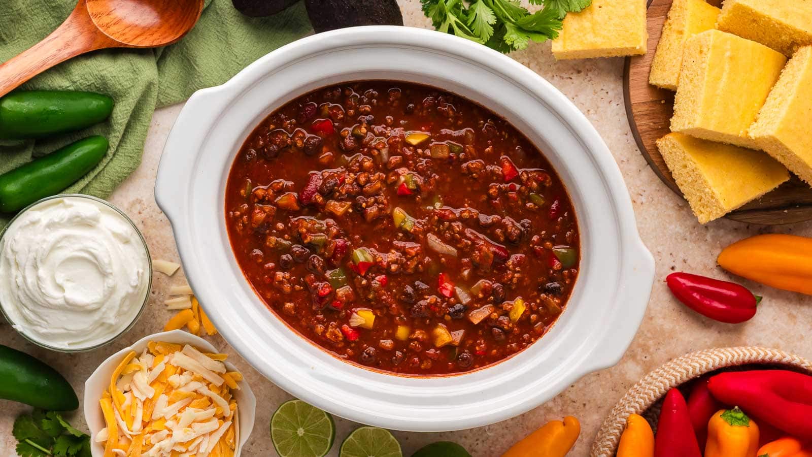Overhead view of slow cooker turkey chili in a white crockpot with sour cream, shredded cheese, cornbread, limes and mini peppers around it by Tessie's Table.