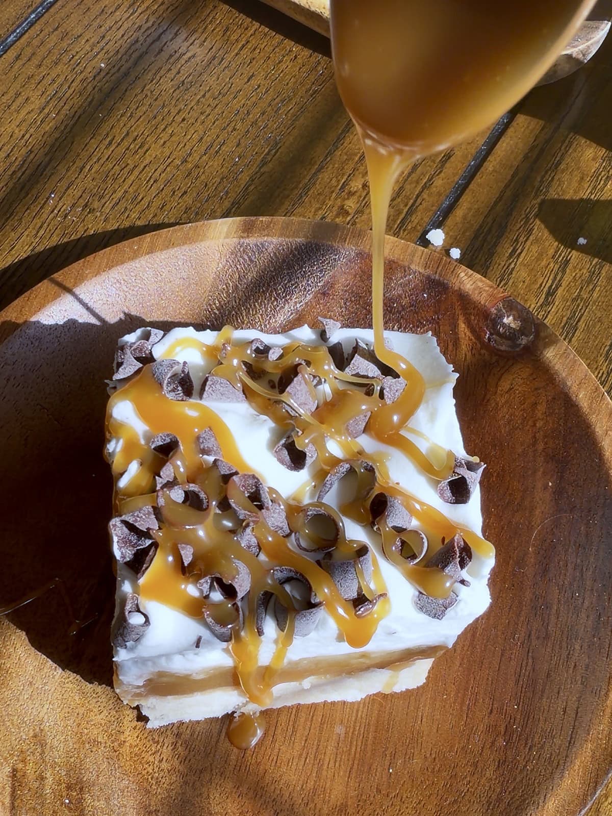 Homemade caramel sauce being drizzled from a spoon over a banoffee shortbread bar topped with whipped cream and chocolate curls by Tessie's Table.