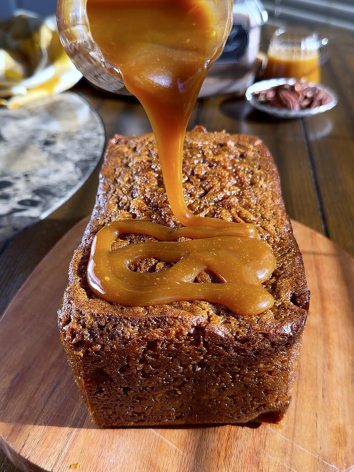 Homemade caramel sauce being poured from a glass jar over a loaf of caramel pecan banana bread by Tessie's Table.