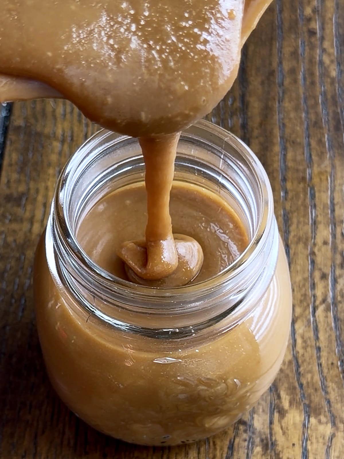 Close up of thick homemade caramel sauce being poured into a glass jar by Tessie's Table.