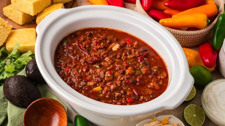 Slow cooker turkey chili with beans in a white crockpot with cornbread, avocado, limes, sour cream and mini peppers around it by Tessie's Table.