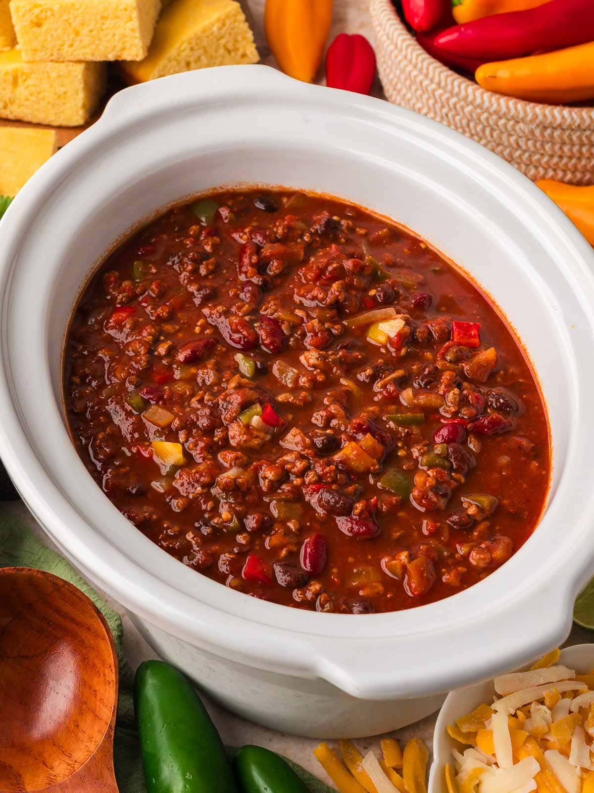 Slow cooker turkey chili with beans in a white crockpot surrounded by cornbread, mini peppers, jalapenos, sour cream and shredded cheese by Tessie's Table.