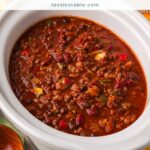 Slow cooker turkey chili with beans in a white crockpot surrounded by cornbread, mini peppers, jalapenos, sour cream and shredded cheese.