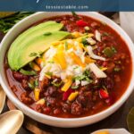 Bowl of turkey chili with beans topped with sliced avocado, sour cream, shredded cheese and cilantro.