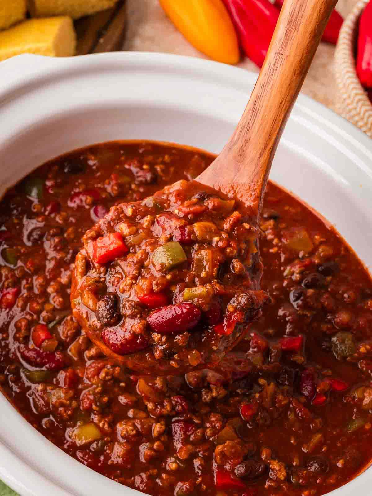 Wooden spoon scooping slow cooker turkey chili with beans and bell peppers from a white crockpot by Tessie's Table.