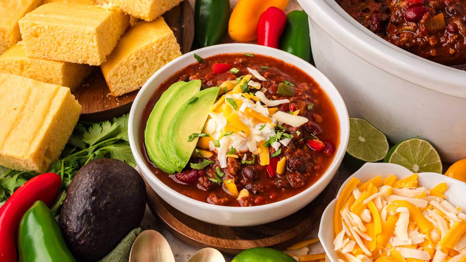 Bowl of slow cooker turkey chili topped with avocado, sour cream, shredded cheese and cilantro next to cornbread, peppers and the slow cooker by Tessie's Table.