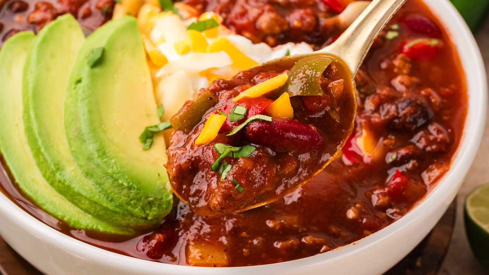 Gold spoon lifting a bite of turkey chili with kidney beans and peppers from a bowl topped with sliced avocado and sour cream by Tessie's Table.